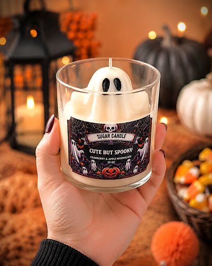 A hand holds a glass jar candle shaped like a white ghost with black eyes. The candle has a Halloween-themed black label reading 'Cute But Spooky' and 'Sugar Candle.