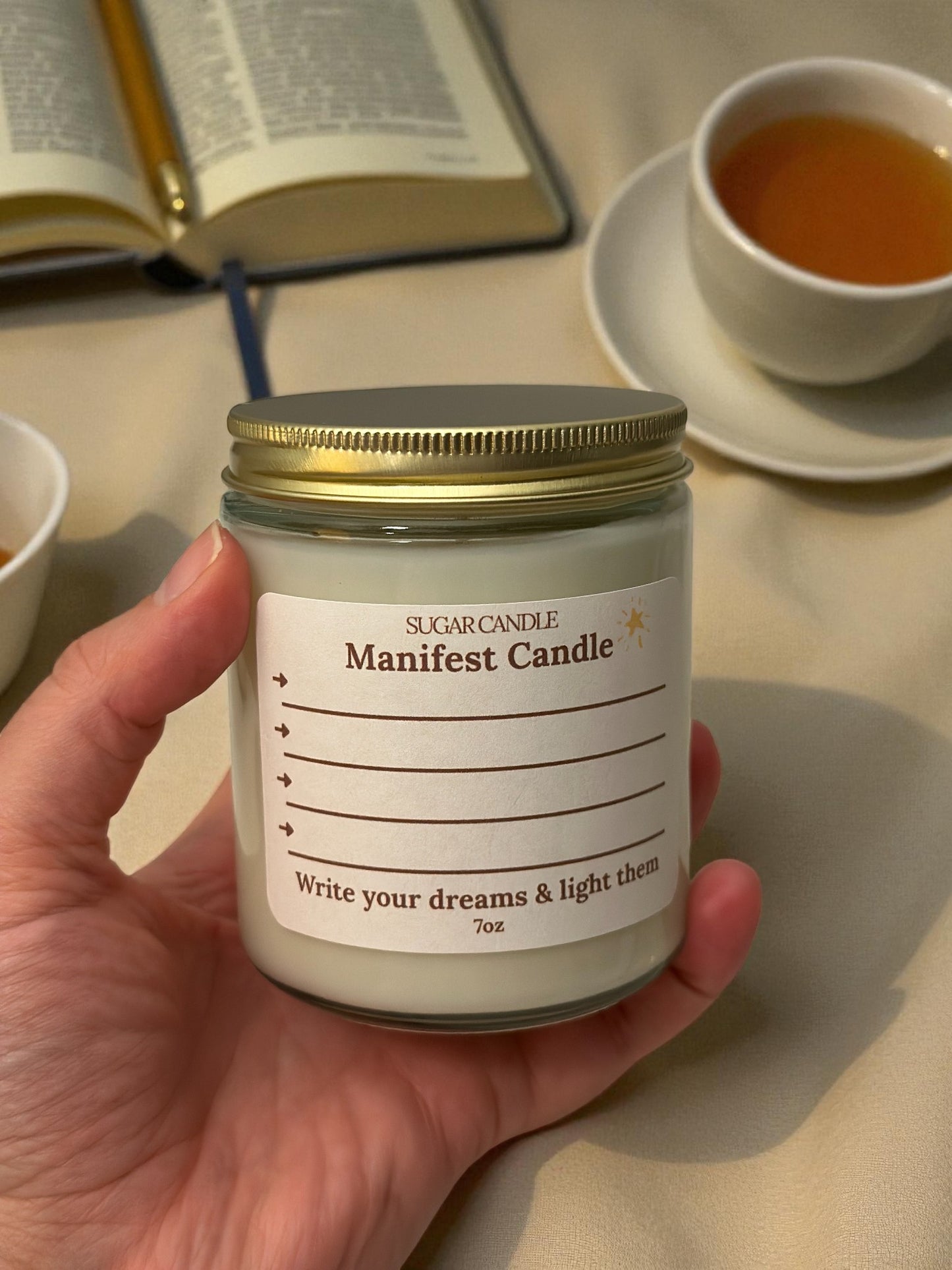 Hand holding a 'Manifest Candle' with an open book and cup of tea in the background
