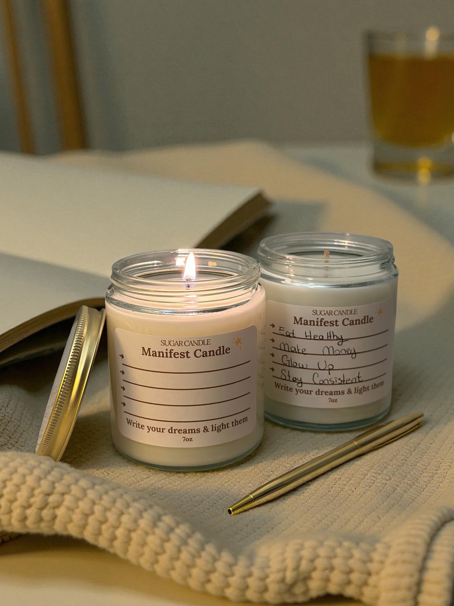 Two lit candles in glass jars with labels on a surface with a book and glass in the background.