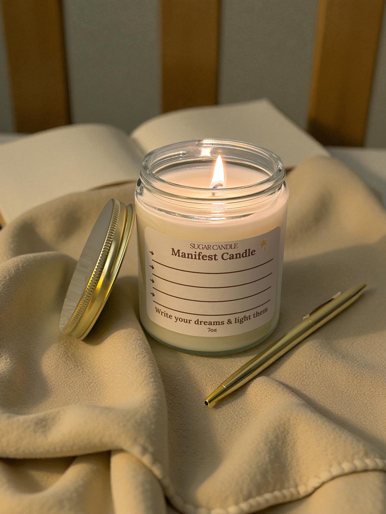 Lit candle in a jar labeled 'Manifest Candle' on a soft fabric surface