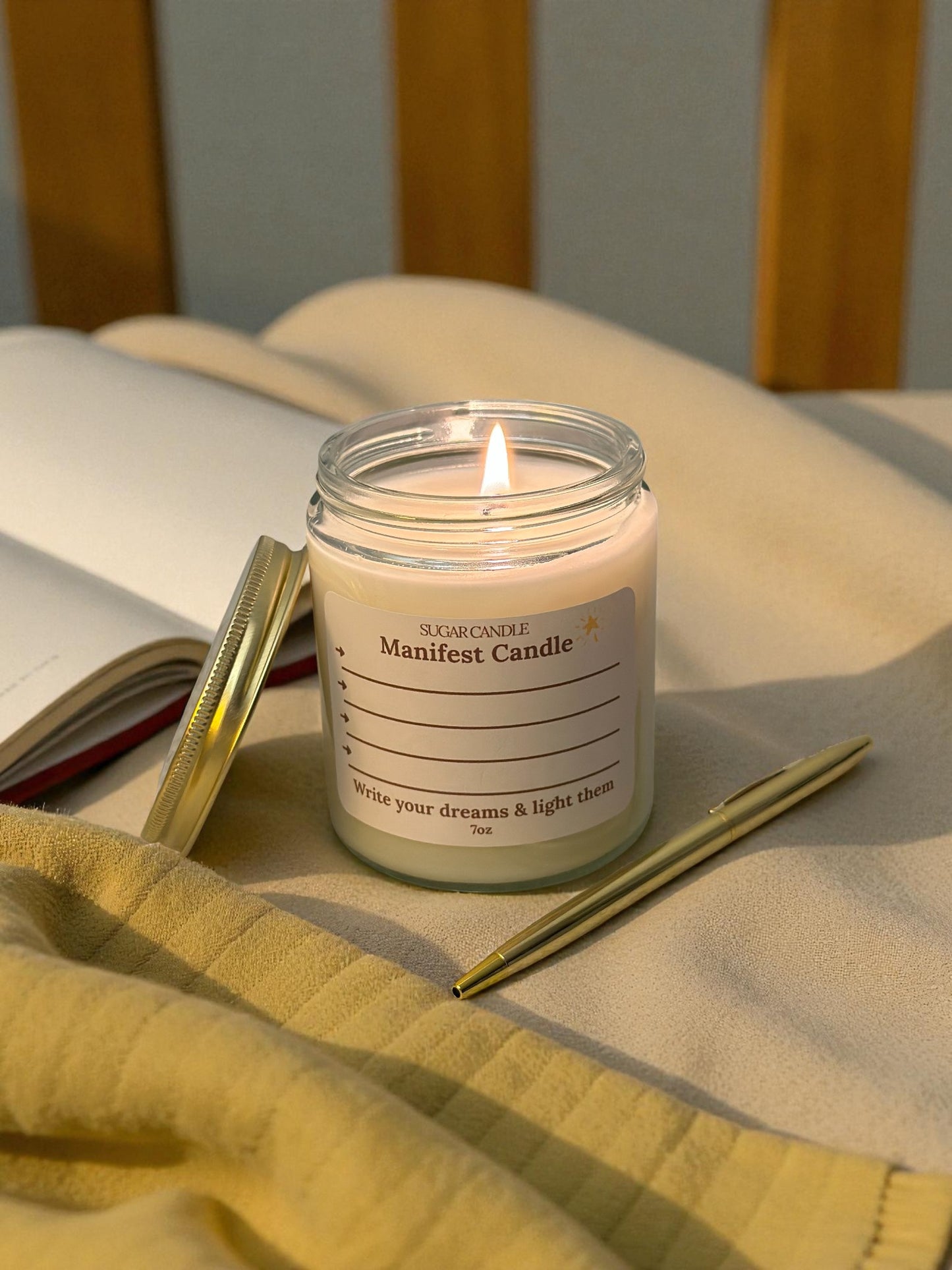 Candle labeled 'Manifest Candle' on a bed with an open book and pen.