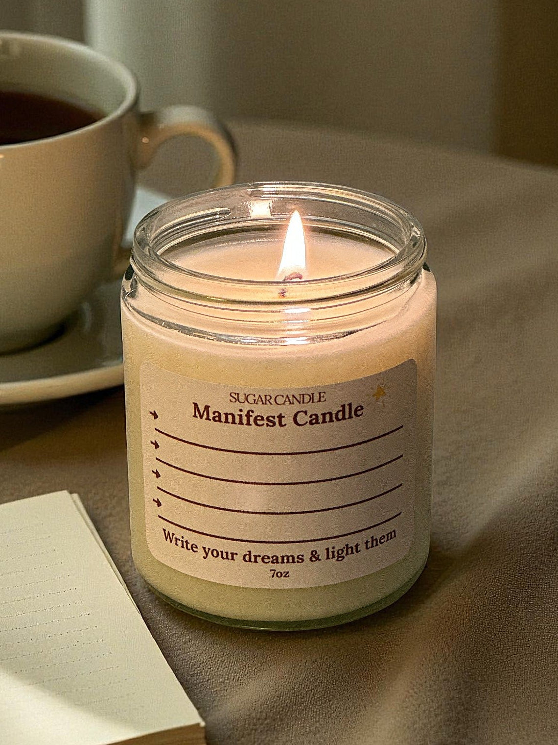 Candle labeled 'Manifest Candle' next to a cup of coffee and notebook on a table.