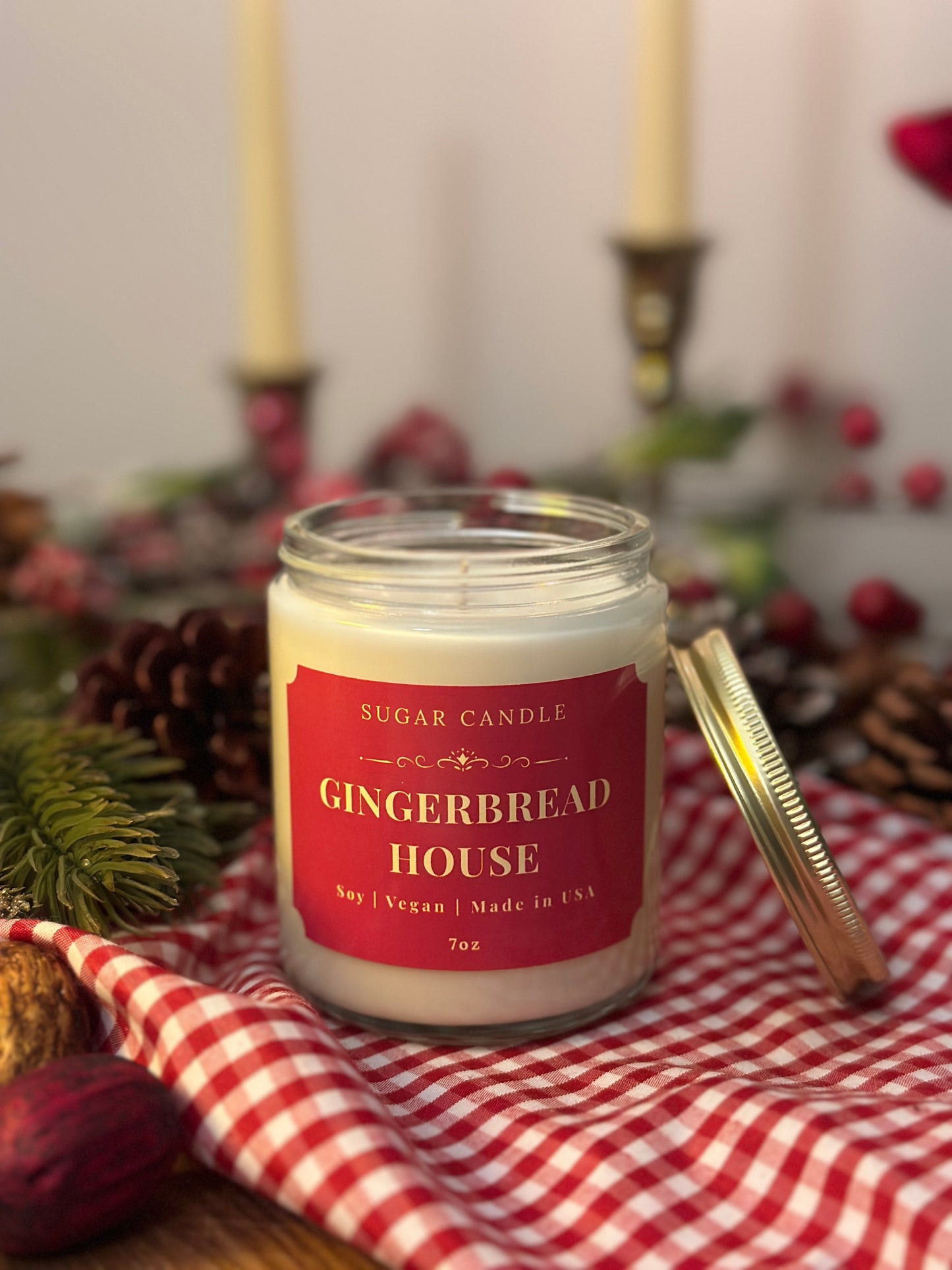 Gingerbread house candle on a red and white checkered cloth with Christmas decorations in the background.