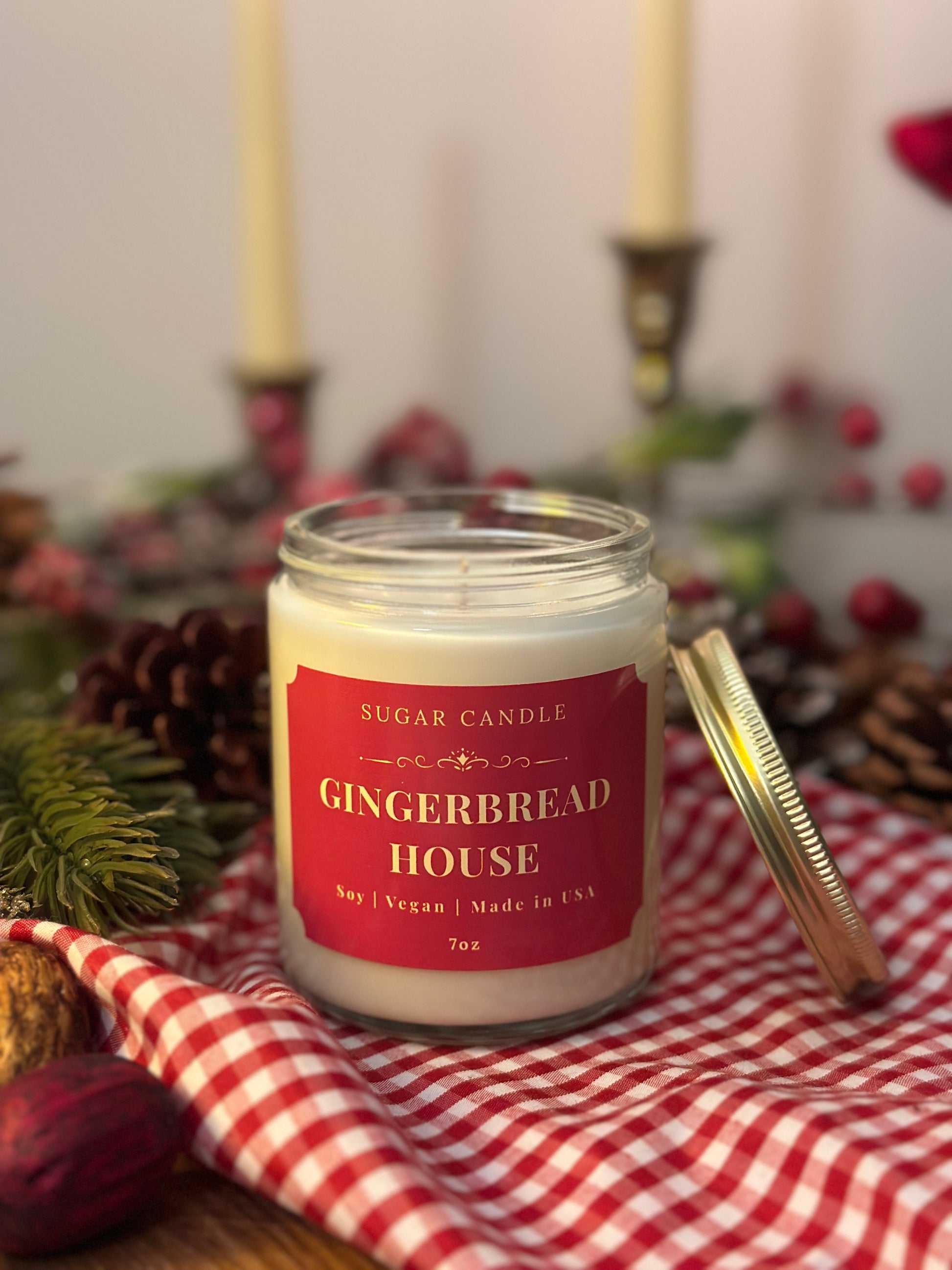 Gingerbread house candle on a red and white checkered cloth with Christmas decorations in the background.