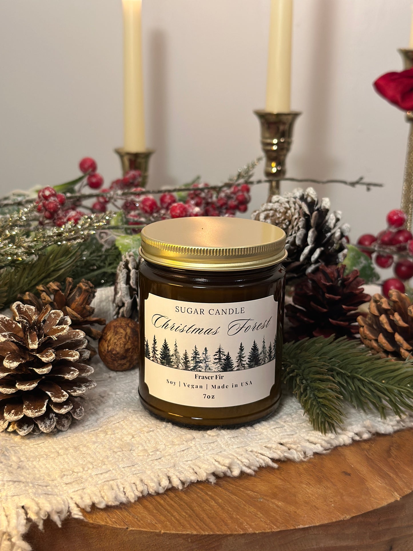 Candle labeled 'Christmas Forest' surrounded by pinecones and evergreen branches on a wooden surface.