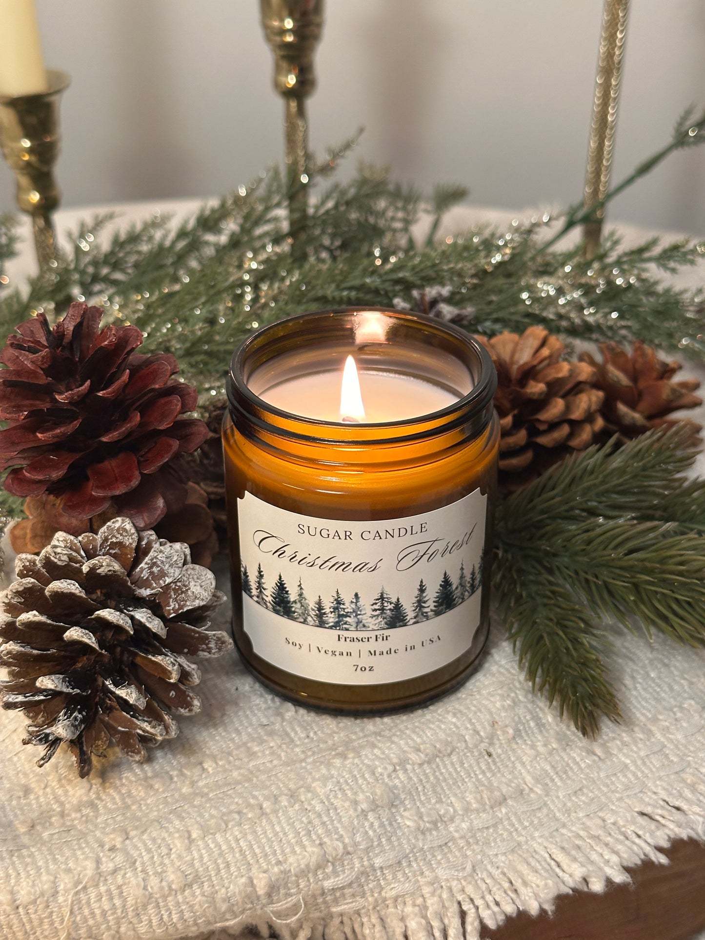 Candle in a glass jar with a label, surrounded by pine cones and branches on a textured surface.