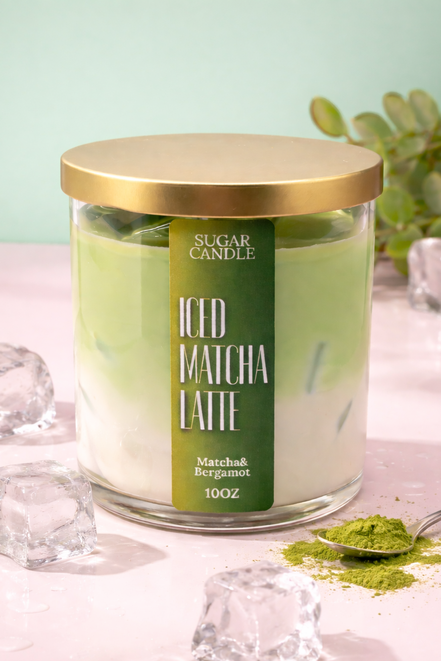 Glass candle jar labeled 'Iced Matcha Latte' with a gold lid, surrounded by ice cubes and matcha powder on a light pink background.