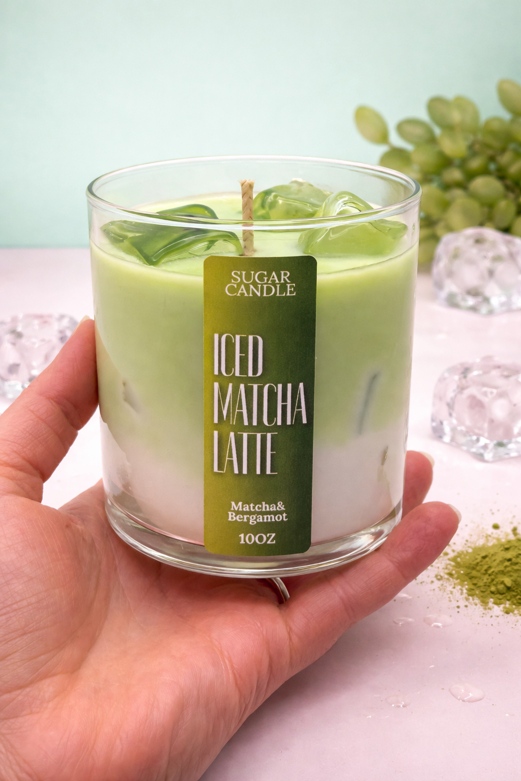 Hand holding a 'Matcha Latte' candle with a green label, surrounded by matcha powder and leaves.